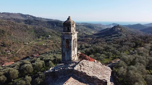 Aerial drone view of bell tower of abandoned church called Chiesa di Sant'Andrea. Bird eye view of old stone roof and bell tower surrounded by olive trees and sea in distance, Imperia, Liguria, Italy