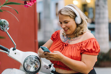 Cheerful woman with headphones using smartphone with scooter on street