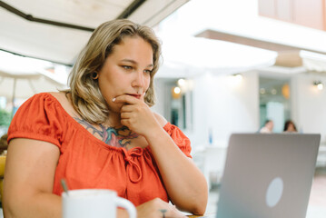 Thoughtful plump woman working on laptop in cafe