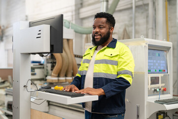 Black man engineer using computer operating CNC machine for cutting wood in furniture factory. Male technical maintenance and programming to machine for making wooden product.
