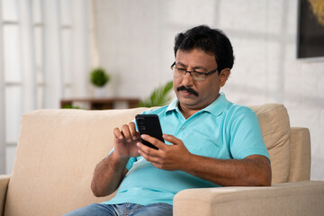 Indian middle aged man with eyeglasses busy using mobile phone while sitting on sofa at home - concept of Digital Engagement, Social Media and Cyberspace