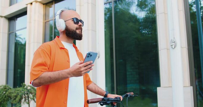 Happy bearded caucasian bearded man in headphone and sunglasses using smartphone while walking with bike at urban area. Commute work sustainable lifestyle in the city.