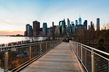 View of the skyscrapers of Lower Manhattan from a walkway path bridge in Brooklyn, lit by sunset.