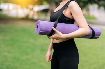 Health-conscious Asian woman stands with a rubber mat in the park, gearing up for sunset exercises. Wellness, activity, and natural beauty.