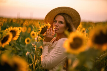 Beautiful middle aged woman looks good in a hat enjoying nature in a field of sunflowers at sunset. Summer. Attractive brunette with long healthy hair.