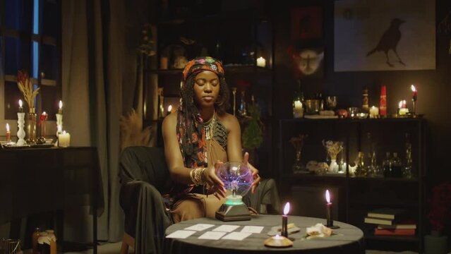 African American concentrated fortune teller predicting future with crystal ball when sitting with closed eyes at table with burning candles in studio