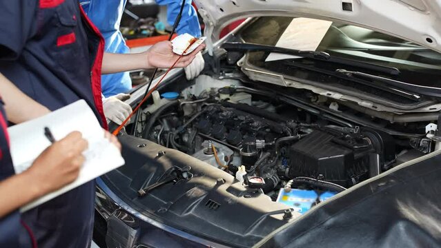 Selective focus, cropped view of a young male mechanic in uniform, standing in front of the car with an open hood, pulling out the dipstick and wipe off the oil with a cloth for checking the oil level