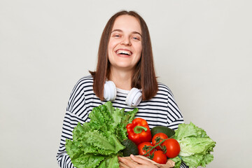 Enjoying a vibrant green diet. Fresh greenery and vegetables. Happy laughing woman wearing striped casual shirt carrying lettuce cabbage pepper avocado isolated over gray background