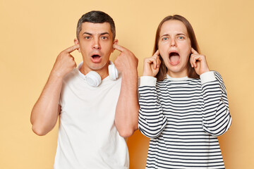 Shocked scared astonished man and woman wearing casual clothing standing isolated over beige background covering ears with fingers do not want to hear something.