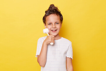 Smiling funny child girl with shampoo on wet hair wearing white T-shirt standing isolated over yellow background taking mother's massager for skin care.