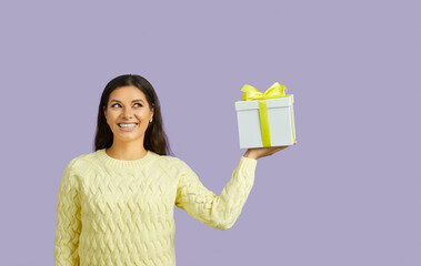 Joyful excited and dreamy woman holding gift box isolated on pastel purple background. Smiling young woman in yellow sweater dreamily looks at copy space. Concept of shopping gifts for the holiday.