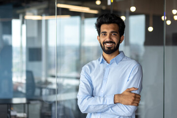 Portrait of young Indian male student, intern, intern standing in blue shirt, arms crossed in office, campus and looking at camera smiling