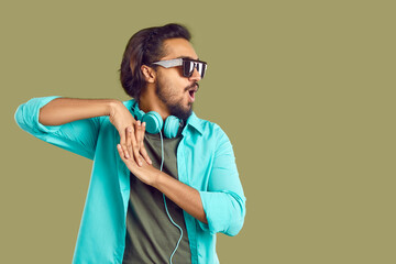 Cheerful indian man with headphones around his neck having fun dancing on khaki background. Bearded man with stereo headphones, in sunglasses and with funny expression makes wave out of his hands.
