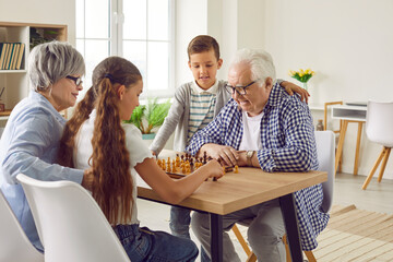 Grandparents and grandchildren spend time together, play chess and have fun. Happy old, retired granddad, grandma and little children sitting at table and playing game of chess. Family leisure concept - obrazy, fototapety, plakaty