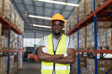 Portrait of African American male warehouse worker wearing safety uniform, helmet standing with crossed arms in the storage warehouse. Man worker working in the warehouse