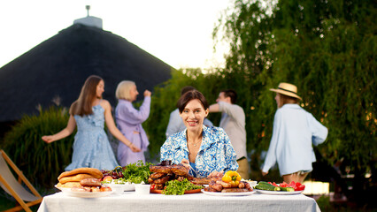 Outdoor dining table with delicious barbecue meats, fresh vegetables and salads. a young woman runs up to the table and takes a steak. Happy people are dancing and having fun in the background.