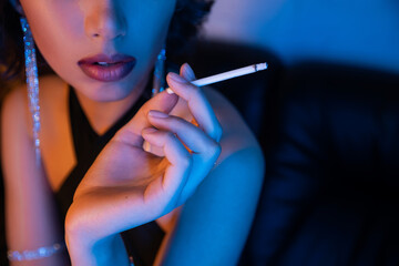 Obraz premium Cropped view of stylish woman in dress holding cigarette while sitting in neon light in night club