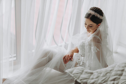 a beautiful girl with a wedding hairstyle and a tiara on her head in a transparent robe is preparing for a wedding in a hotel with a royal interior. Long veil, natural light, portrait photo