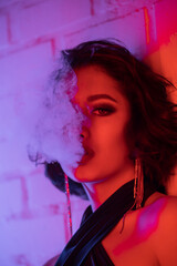 Portrait of trendy young asian woman with makeup exhaling smoke in neon light in night club