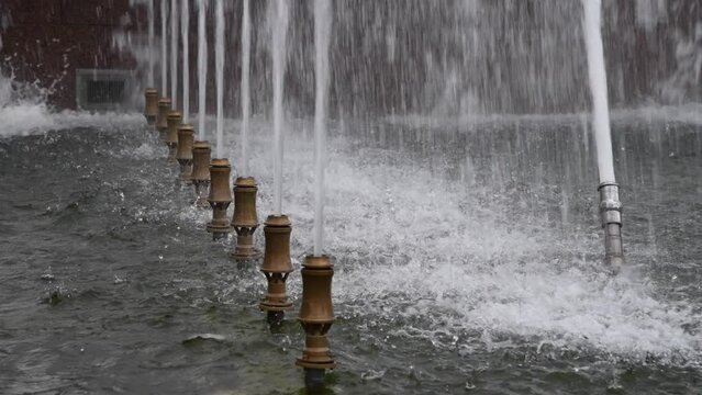 Close-up view of working fountain jet nozzles with splashing water on city square in a cloudy summer day. Real time handheld video. Soft focus. Urban decoration theme.