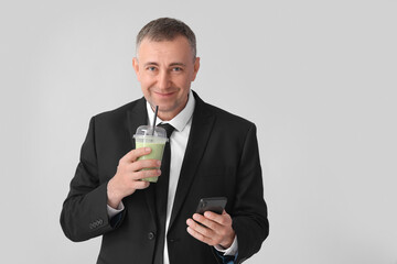 Mature businessman with mobile phone drinking vegetable smoothie on light background