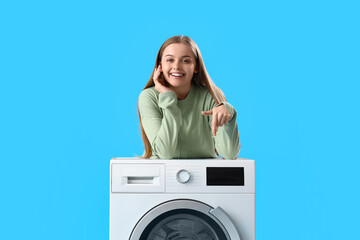 Pretty young woman leaning on washing machine and pointing at it on blue background