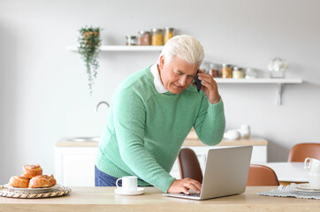 Senior man with laptop talking by mobile phone in kitchen