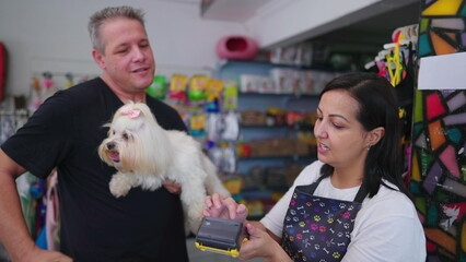 Customer paying for service with contactless credit card on payment machine standing inside Pet Shop holding small dog. Happy client pays for grooming procedure