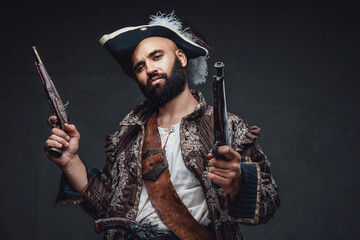 A pirate with a black beard holding two muskets, dressed in a vest and hat, against a dark textured...