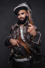 A pirate with a black beard holding two muskets, dressed in a vest and hat, against a dark textured wall