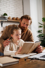 Bearded father hugging daughter reading book near laptop and notebook during e-learning at home