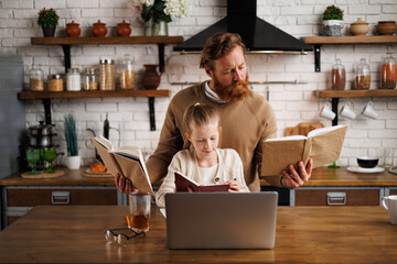 Preadolescent girl holding notebook near bearded dad with books and laptop on table in kitchen at...
