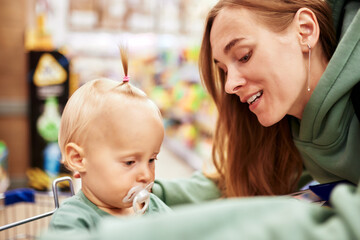 Happy young mother with her cute baby in shopping trolley walking around toy store in shopping mall. Mom and little blonde daughter choosing toys and having fun. Funny family weekend, happy childhood.