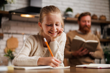 Smiling preadolescent kid writing on notebook near blurred father with book during homeschooling in...