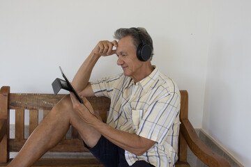 Mature Man outside sitting on a bench watching a show on his iPad Smart Tablet wearing headphones