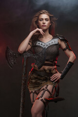 Fototapeta premium A gorgeous Viking girl dressed in chainmail top and fur skirt posing with a two-handed axe, illuminated by red rim light against a textured grey wall