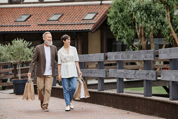 Fototapeta na wymiar positive senior couple walking with shopping bags, elderly man hugging woman, walking together