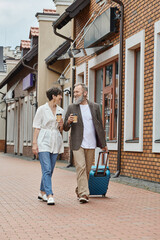 Fototapeta na wymiar happy elderly couple, man and woman walking with coffee to go and luggage on street, urban lifestyle