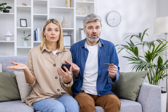 Portrait Of Disappointed Cheated Family, Man And Woman Mature Couple Sitting Sad On Sofa And Looking At Camera, Using Bank Credit Card And Phone For Online Shopping On The Internet