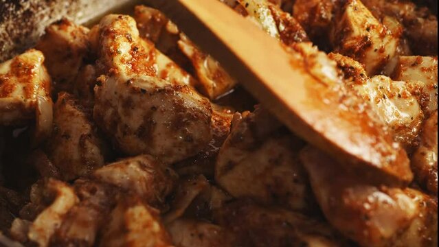 Pieces of chicken fried in a cast-iron skillet in oil with onions and spices are mixed with a wooden spatula