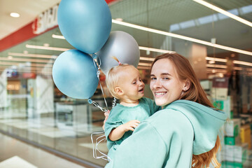Young attractive mother with baby in her arms and colored balloons. Happy smiling mom and her little daughter in same green clothes walk around the mall and have fun. Family weekend in shopping mall.
