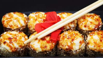 Baked sushi roll in soy sauce with ginger and chopsticks on a black background.