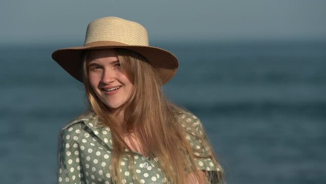 Happy young blonde woman smiling and playfully looking at camera. Cheerful Caucasian female in straw hat and light green polka dot dress on background of sea on summer day. Cinematic shot, slow motion