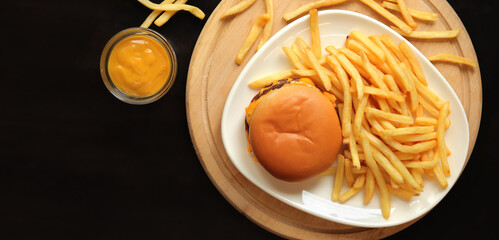 Plate with cheeseburger and french fries, top view. Delicious french fries on a white dish, on a cutting board with mustard, dark background. Copy space for your text