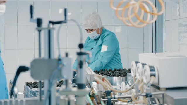 A woman in uniform wearing a mask works on a vial filling line. Production of medical dietary supplements. Distribution of caps. Pharmaceutical production line at pharma factory