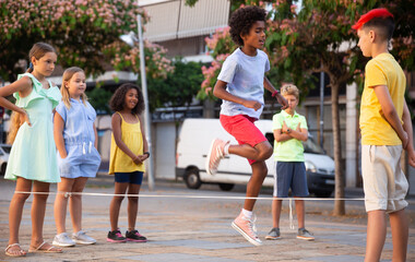 Happy preteen kids of different nationalities skipping on chinese jump rope in courtyard on summer day..