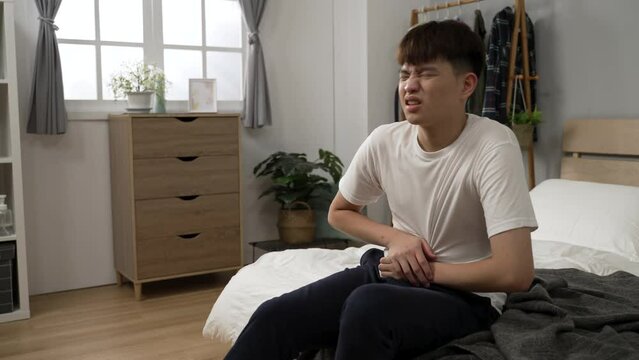 ill asian young man suffering diarrhea is pressing his lower abdomen and moaning with a painful look on face at bedside in the morning in the bedroom at home