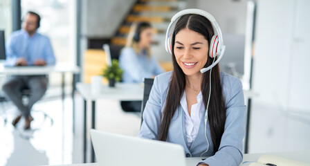 Portrait of friendly female customer support representative in headset with microphone consulting client while using laptop in customer support service office.