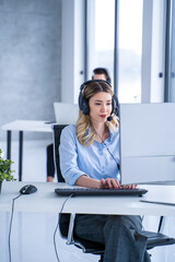 Young blonde woman in headset, sitting in office, working with her colleagues in customer support...