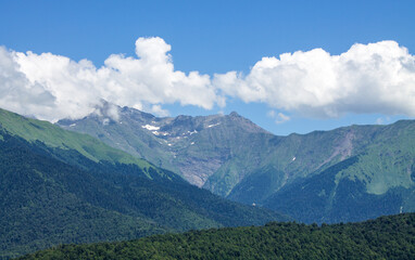 Beautiful panoramic landscape - peaks of high mountains between green trees and blue sky with white clouds on Krasnaya Polyana in Krasnodar krai Russia